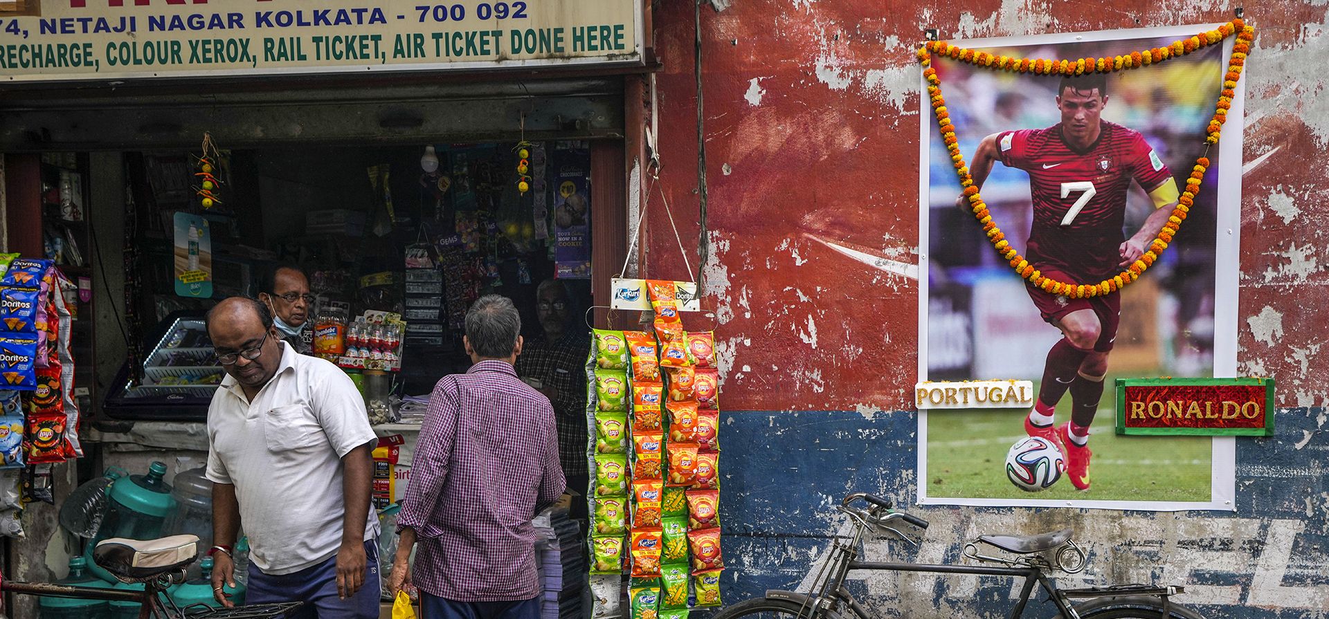 La gente compra comestibles en una tienda junto a una fotografía del portugués Cristiano Ronaldo, adornada con guirnaldas y colocada por fanáticos, para conmemorar la Copa del Mundo de fútbol en Calcuta, India, el jueves 8 de diciembre de 2022. (AP Foto/Bikas Das)