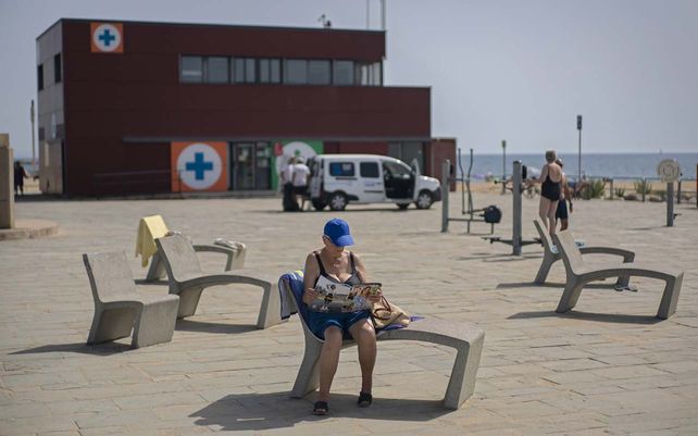 Una mujer lee una revista en un paseo marítimo frente a un edificio de la Cruz Roja en Barcelona. En España