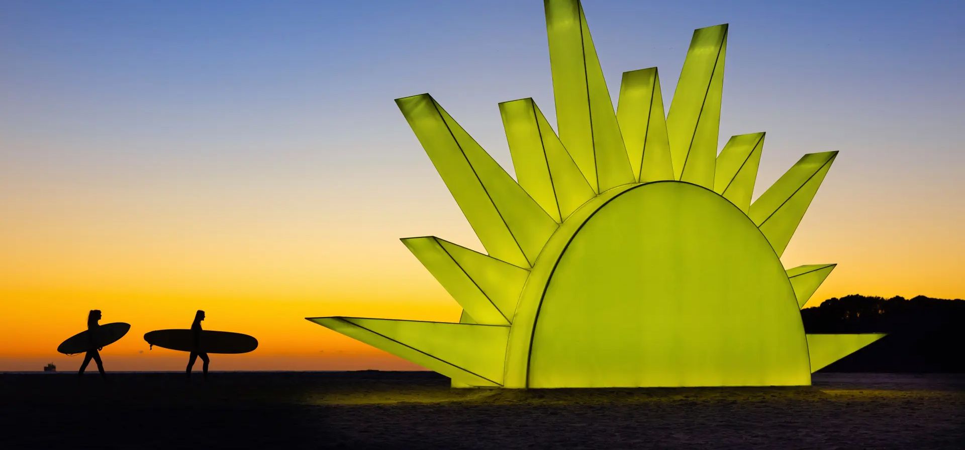 Isla Norte, Nueva Zelanda. Los surfistas pasan junto a una escultura gigante del sol al amanecer en la playa del Monte Maunganui, Second Sun es una lámpara de terapia de luz iluminada por más de 8.000 bombillas. La obra de arte de 18 metros de ancho emite una luz diseñada para levantar el estado de ánimo durante los oscuros meses de invierno. Fotografía: Brett Phibbs/AP