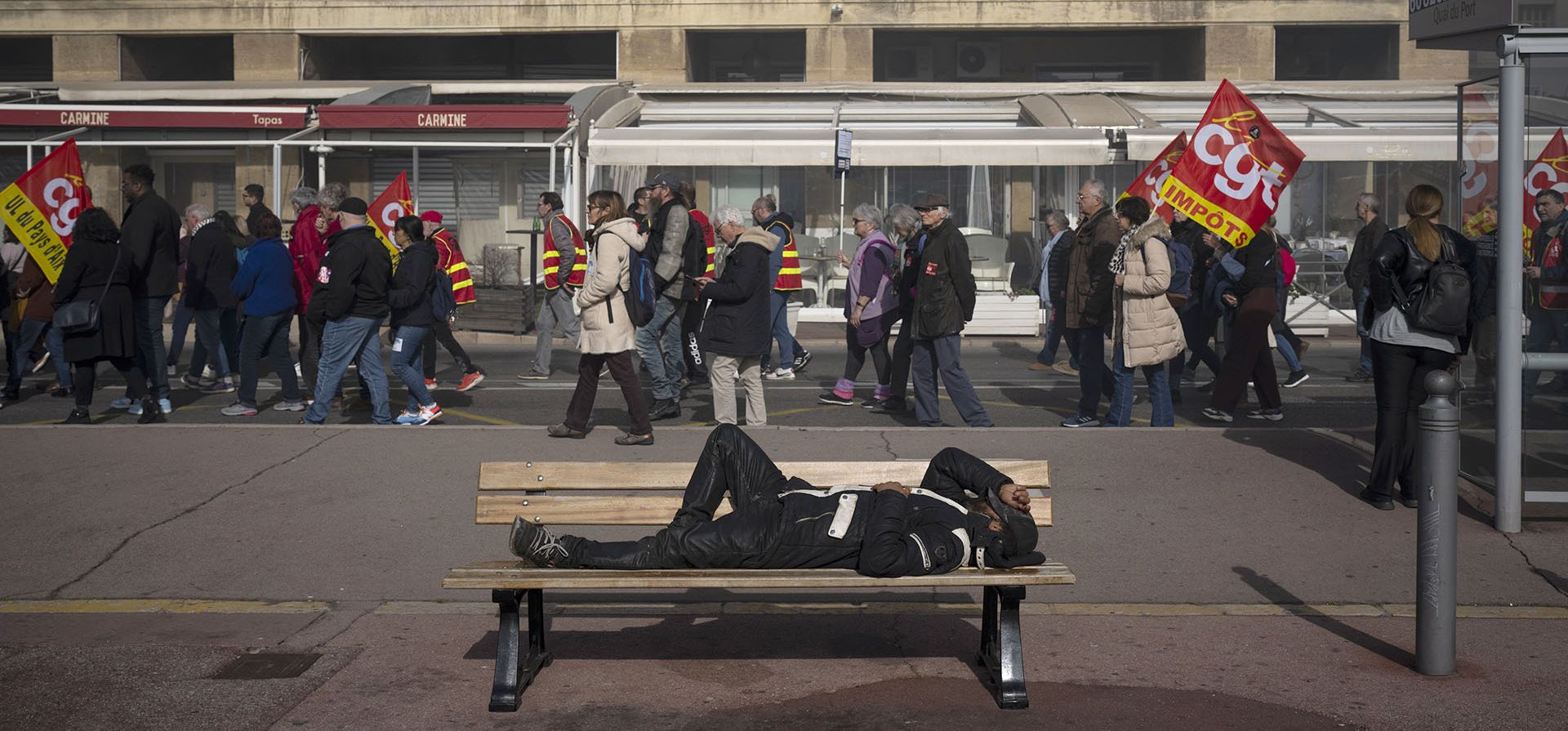 Un hombre duerme en un banco mientras pasa una manifestación en Marsella, sur de Francia, el jueves 16 de febrero de 2023. Francia se prepara para el quinto día de huelgas y protestas nacionales el jueves contra la reforma de las pensiones, que es la política insignia del presidente Emmanuel Macron. segundo período. (Foto AP/Daniel Cole)