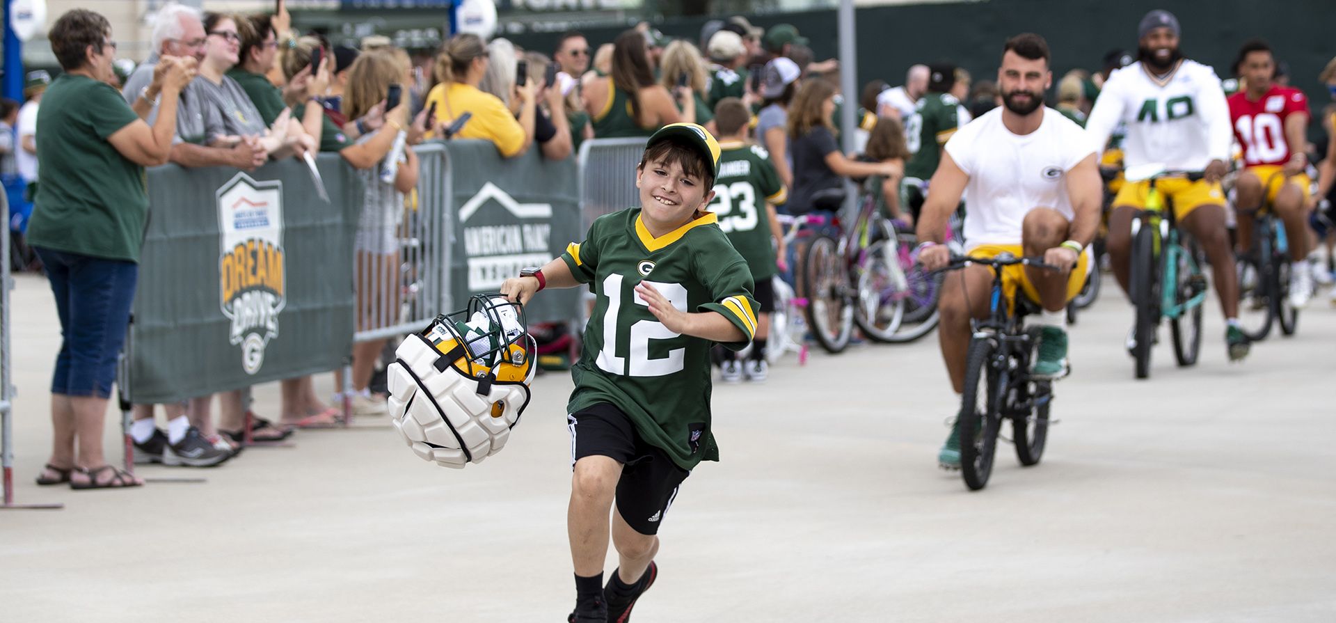 El ala cerrada de los Green Bay Packers, Josiah Deguara, tercero desde la derecha, monta la bicicleta de un joven aficionado mientras el aficionado sale corriendo con el casco de Deguara durante el campamento de entrenamiento de fútbol americano de la NFL del equipo, en Green Bay, Wisconsin.