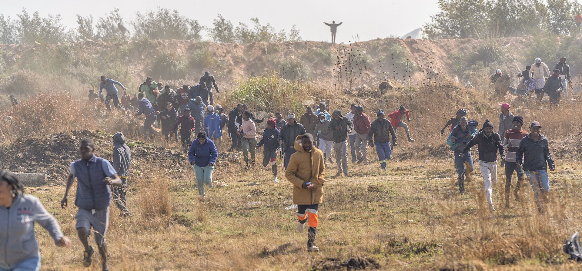 La gente huye de los gases lacrimógenos y las granadas lanzadas desde un helicóptero de la policía en la ciudad sudafricana de Krugersdorp el jueves 4 de agosto de 2022. Los miembros de la comunidad atacaron a presuntos mineros ilegales e incendiaron sus campamentos el jueves en un estallido de ira tras el ataque por las presuntas violaciones en grupo de ocho mujeres por mineros la semana pasada.
