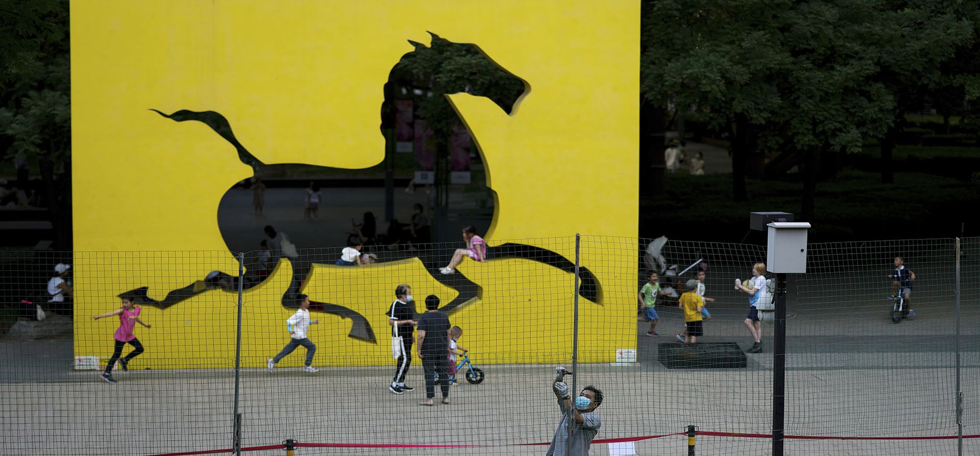 Los niños juegan cerca de una instalación de arte en un complejo de edificios residenciales mientras un trabajador arregla una barricada para evitar que los residentes se reúnan en un espacio verde debido a las restricciones de COVID-19 en Beijing, el jueves 26 de mayo de 2022.