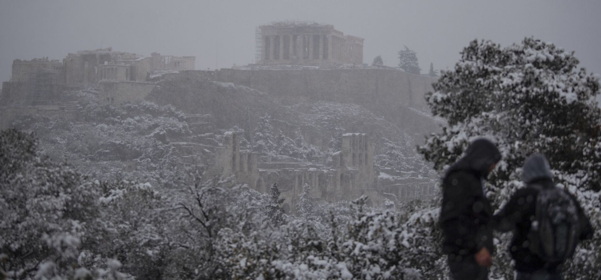 Dos personas caminan frente a la antigua colina de la Acrópolis durante la nevada, en el centro de Atenas. Una tormenta de nieve azotó la capital griega de Atenas, entorpeciendo el tráfico, forzando la cancelación de vuelos y suspendiendo las vacunas contra el coronavirus en la región más amplia de la capital griega.