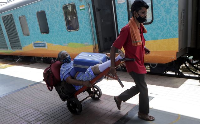 Un trabajador de ferrocarril transporta en un carrito de equipaje a una pasajera anciana que tiene dificultades para caminar mientras la ayuda a abordar un tren en Mumbai