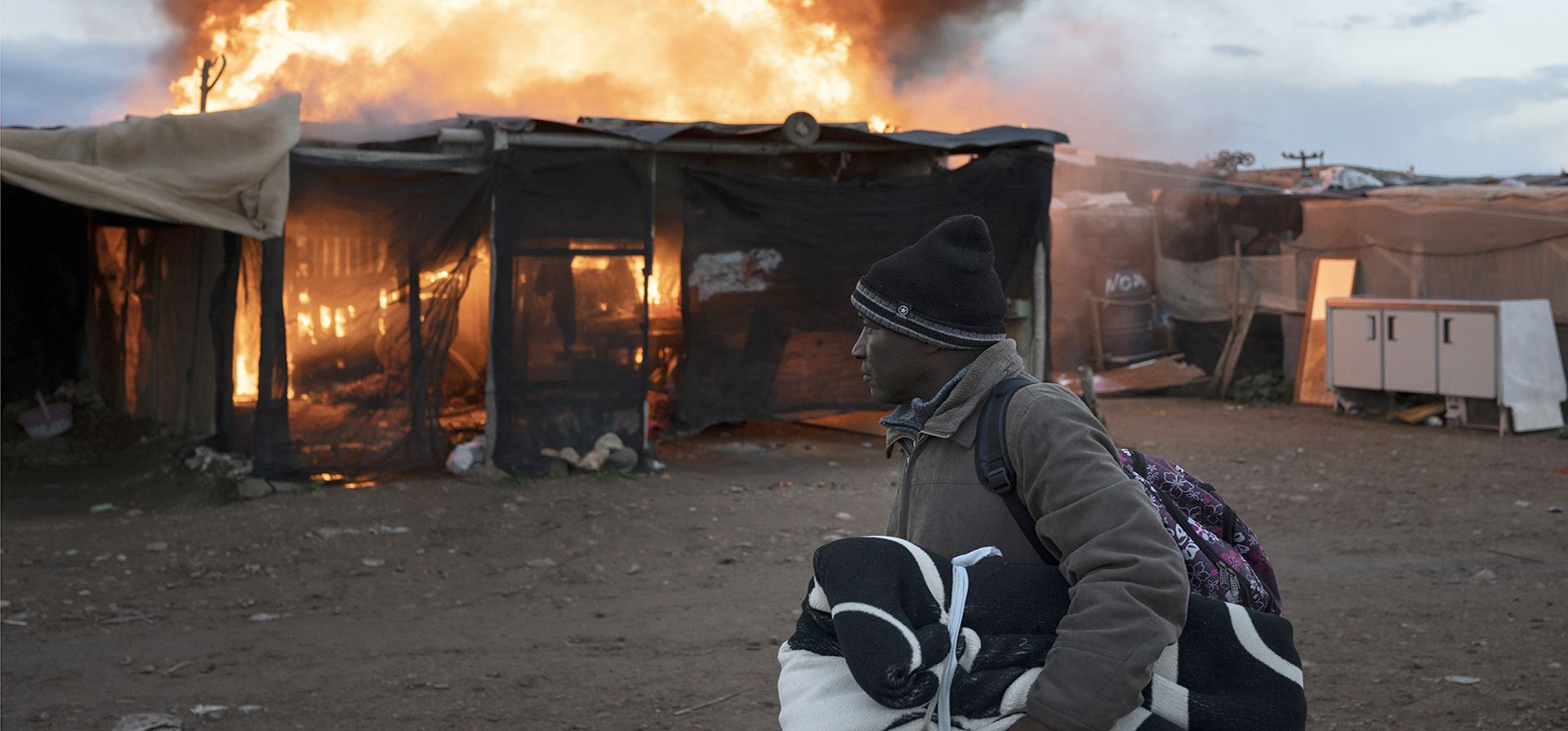 Un hombre que lleva sus pertenencias pasa junto a chozas en llamas en un campamento en Almería, España, el lunes 30 de enero de 2023. Bomberos extinguieron un incendio en un barrio marginal habitado por trabajadores migrantes, cerca del pueblo de Níjar, en el sur de España. (Foto AP/Santi Donaire)