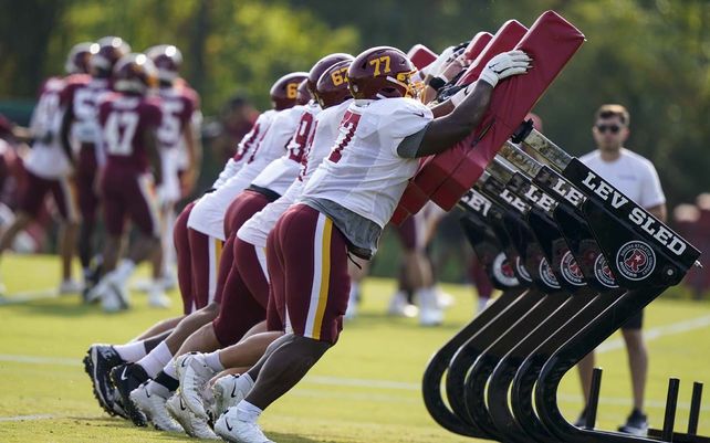 El tackle ofensivo del Washington Football Team Saahdiq Charles (77) y otros miembros de la línea ofensiva trabajan contra un sled de entrenamiento durante la práctica en las instalaciones de práctica de fútbol americano de la NFL