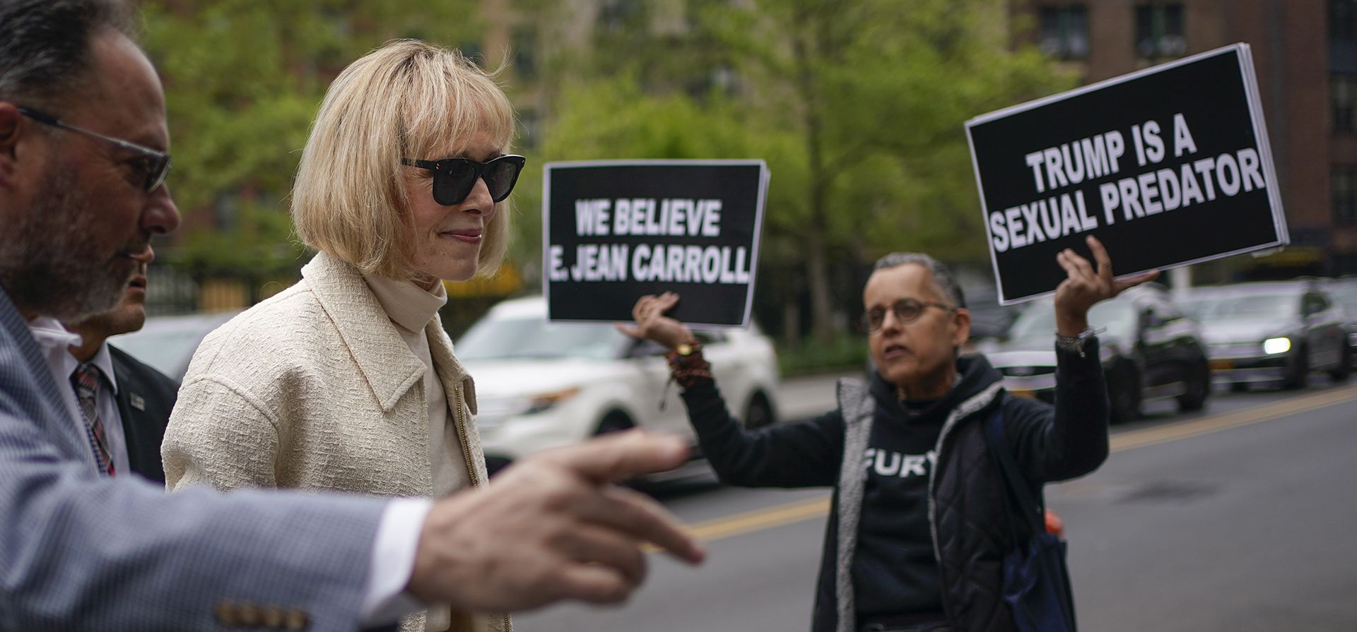 Mientras un manifestante sostiene carteles, E. Jean Carroll llega a la corte federal en Nueva York el jueves 27 de abril de 2023. Carroll comenzó a testificar el miércoles en el juicio de su demanda federal. La escritora le dijo a un jurado que Donald Trump la violó después de que ella lo acompañó al probador de una tienda departamental de lujo en 1996. (AP Photo/Seth Wenig)