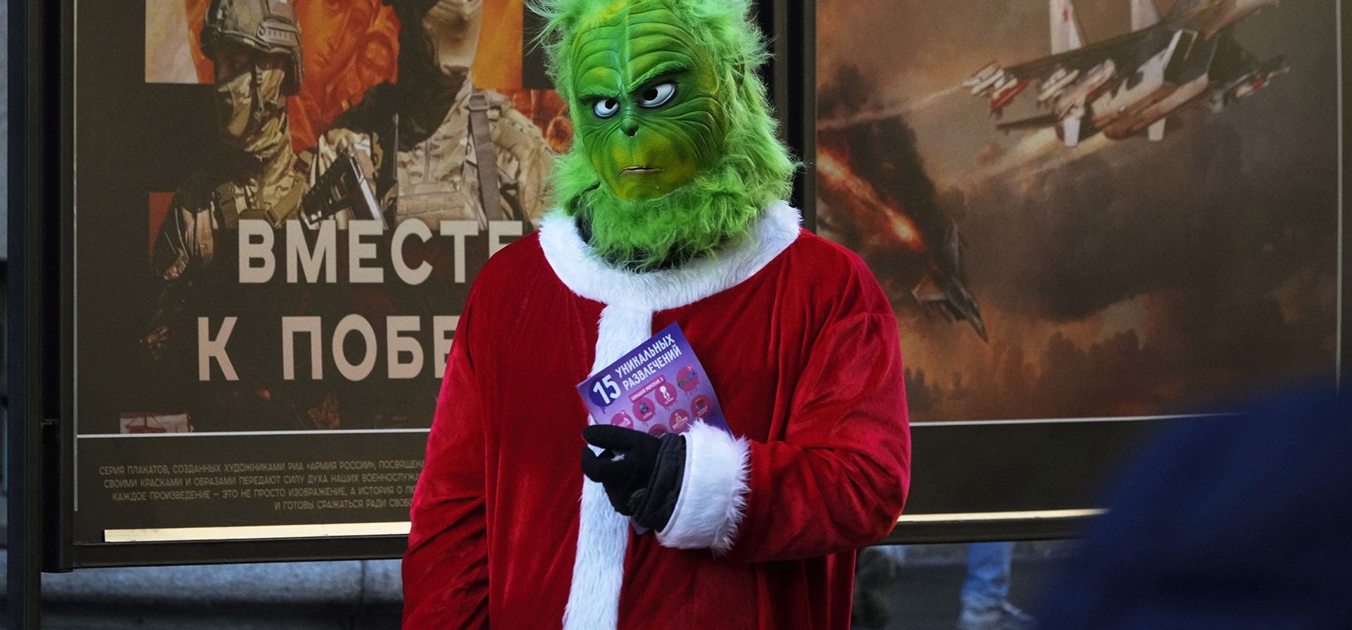 Un artista callejero disfrazado de Grinch invita a la gente a visitar un parque de diversiones, mientras se encuentra junto a una exposición callejera de carteles militares llamada 