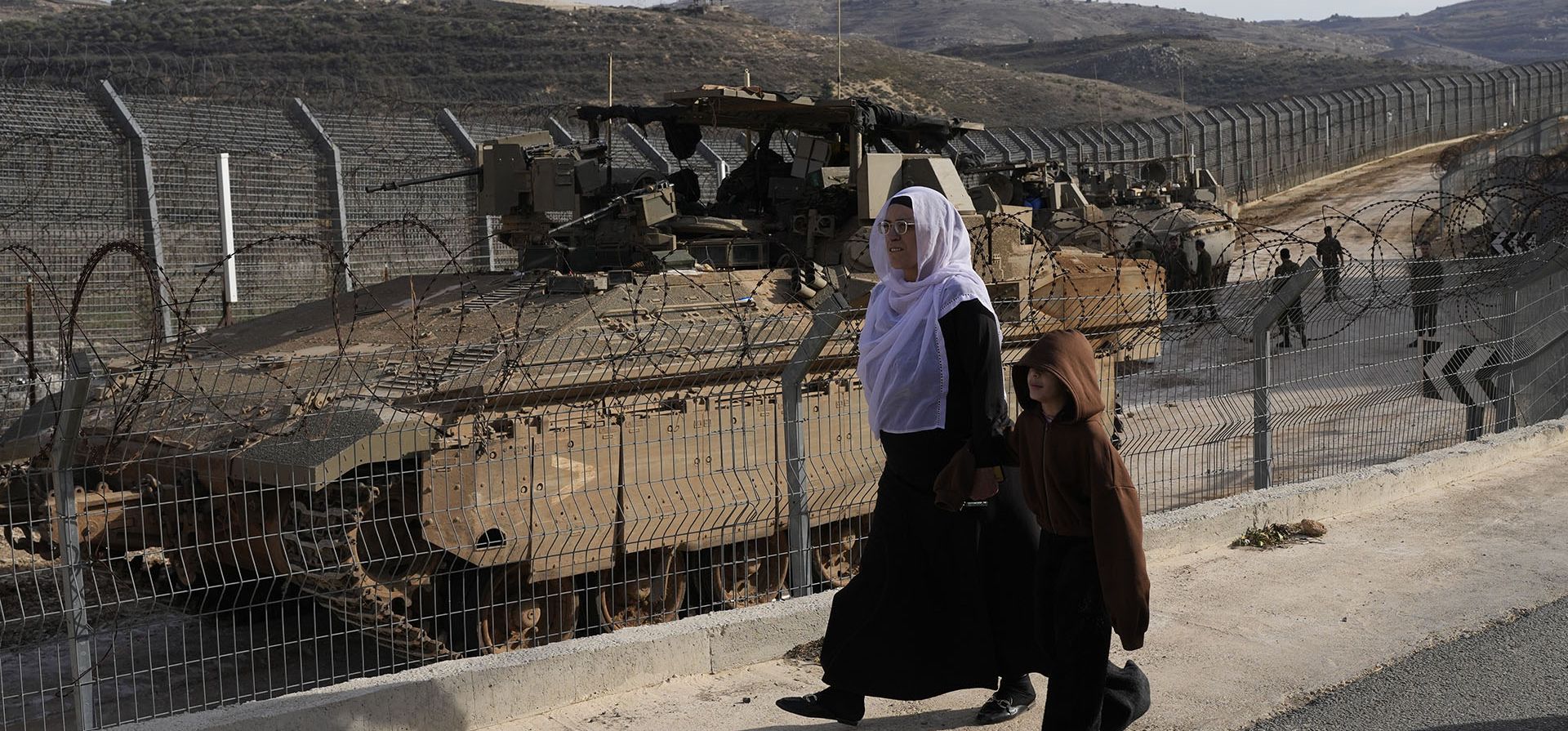 Residentes caminan junto a vehículos blindados israelíes estacionados cerca de la llamada Línea Alfa que separa los Altos del Golán anexados por Israel de Siria, en la ciudad de Majdal Shams, el lunes 9 de diciembre de 2024. (Foto AP/Matias Delacroix)