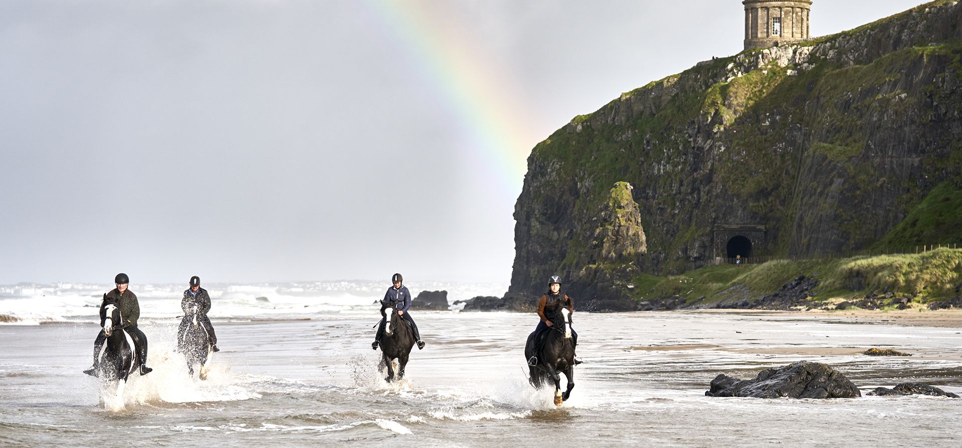 Visitantes disfrutan del paseo a caballo por la ruta costera de Causeway con vistas al templo Mussenden en la playa Downhill, con un arcoíris como parte de la experiencia Coastal Canters Embrace a Giant Spirit. Foto: AP Visitantes disfrutan del paseo a caballo por la ruta costera de Causeway con vistas al templo Mussenden en la playa Downhill, con un arcoíris como parte de la experiencia Coastal Canters Embrace a Giant Spirit. Foto: AP