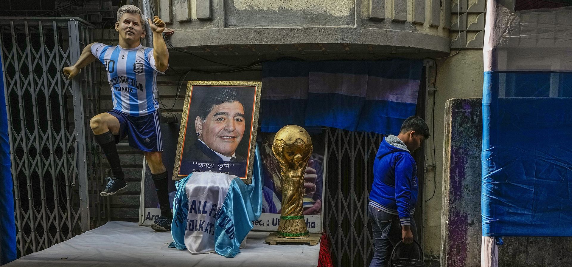 Un niño lleva agua potable frente a un retrato de la leyenda del fútbol argentino Maradona junto a una réplica de la Copa del Mundo exhibida con motivo de la Copa del Mundo de fútbol en curso en Calcuta, India, el jueves 8 de diciembre de 2022. (Foto AP/Bikas Das)