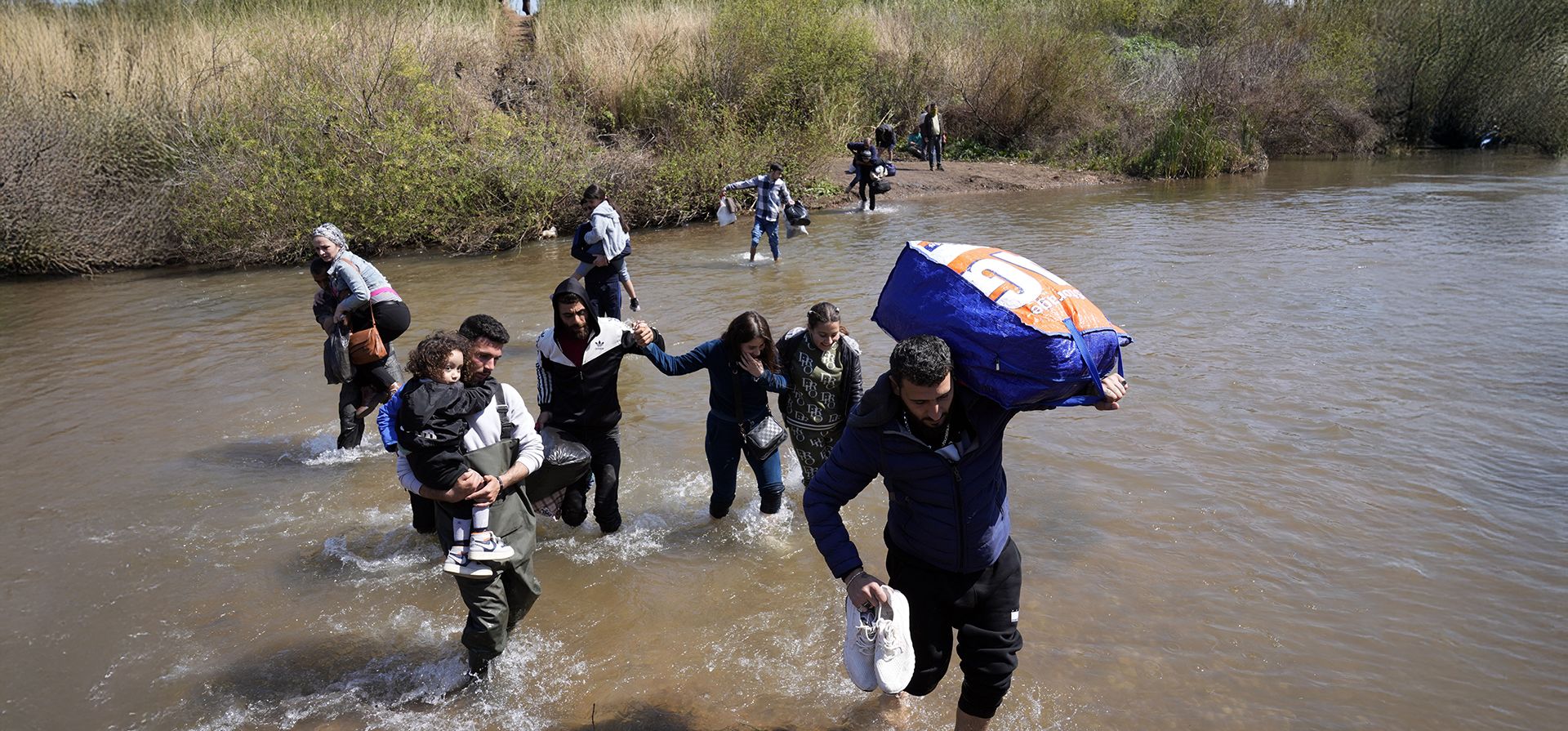 Familias sirias que huyeron de los enfrentamientos en Siria sostienen sus maletas mientras cruzan un río que marca la frontera entre Siria y el norte del Líbano, cerca de la aldea de Heker al-Daher, en la provincia de Akkar, Líbano, el martes 11 de marzo de 2025. (Foto AP/Hussein Malla)