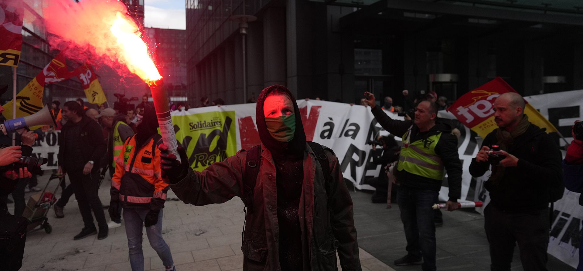 Manifestantes sostienen bengalas y pancartas en protesta contra las reformas de las pensiones frente al edificio Euronext Paris en el distrito comercial de La Defense en París, el jueves 20 de abril de 2023. Activistas sindicales realizan acciones dispersas para presionar al gobierno de Francia para que elimine la nueva ley que eleva la edad de jubilación. (Foto AP/Thibault Camus)