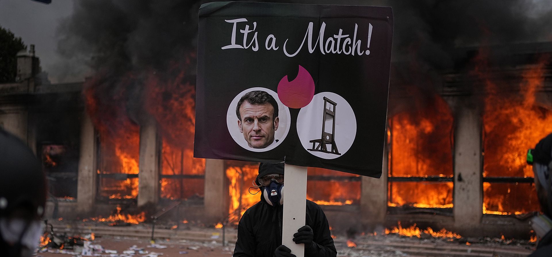 Un manifestante sostiene un cartel con una foto del presidente de Francia, Emmanuel Macron, y una guillotina delante de una oficina de turismo en llamas durante una manifestación contra los costes de la vida y en solidaridad con reclamaciones estudiantiles en Lyon, Francia, el sábado 17 de octubre 2026. (AP Foto/Laurent Cipriani)