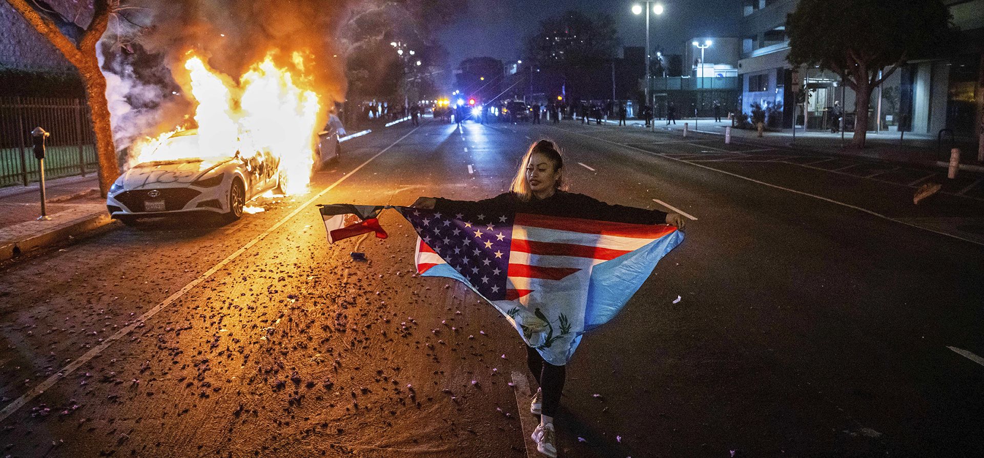 Una persona con varias banderas camina junto a un coche en llamas durante las protestas por las redadas de inmigración de la administración Trump en Los Ángeles, el lunes 9 de junio de 2025. (Foto AP/Ethan Swope) Una persona con varias banderas camina junto a un coche en llamas durante las protestas por las redadas de inmigración de la administración Trump en Los Ángeles, el lunes 9 de junio de 2025. (Foto AP/Ethan Swope)