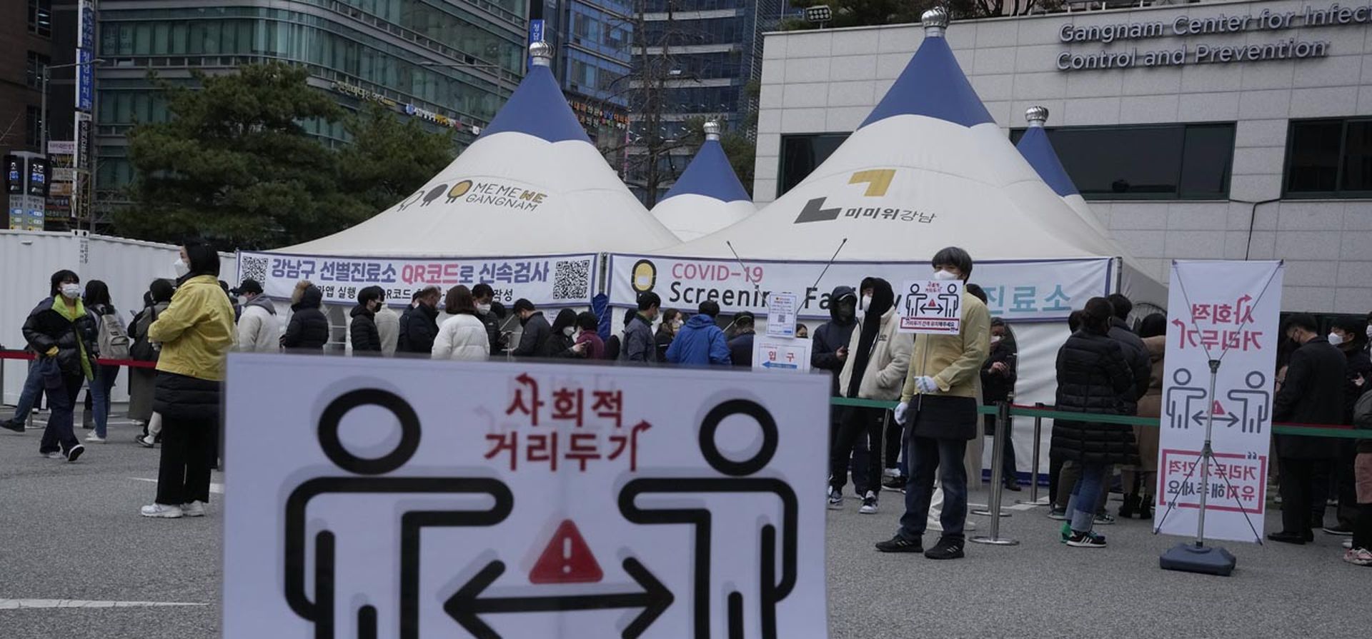 Señales de distanciamiento social sobre las precauciones contra el coronavirus se muestran mientras las personas hacen fila para las pruebas de coronavirus en un centro de salud pública en Seúl, Corea del Sur, el jueves 9 de diciembre de 2021. Las nuevas infecciones por coronavirus en Corea del Sur han estado aumentando y batiendo récords diario.
