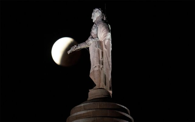 La sombra de la tierra cubre la luna llena durante un eclipse lunar parcial visible cerca de una estatua de George Washington en lo alto del Monumento en Baltimore