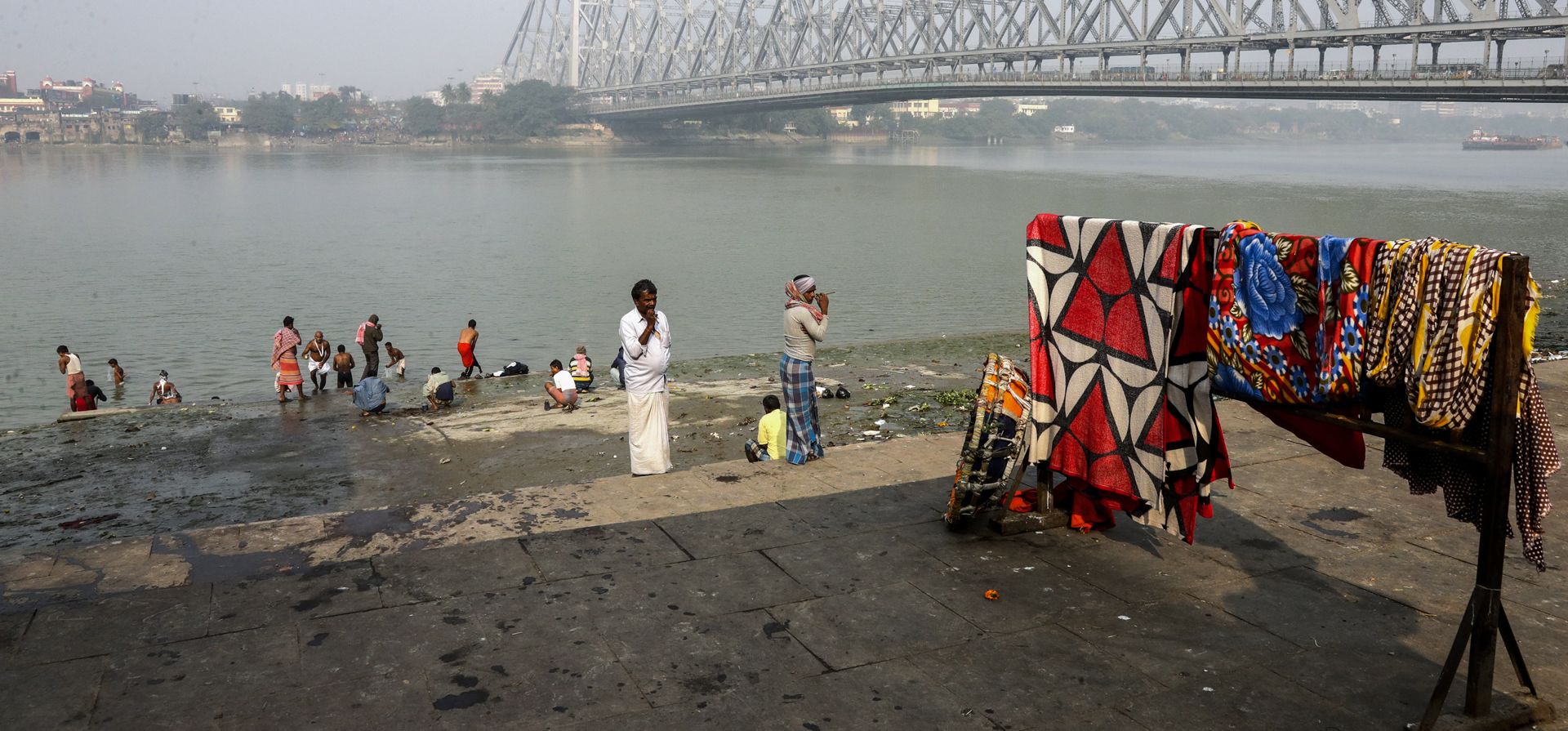 La gente extiende ropa para secarse mientras otras personas se bañan en el río Hooghly junto al puente Howrah en Calcuta, India, el lunes 17 de enero de 2022.