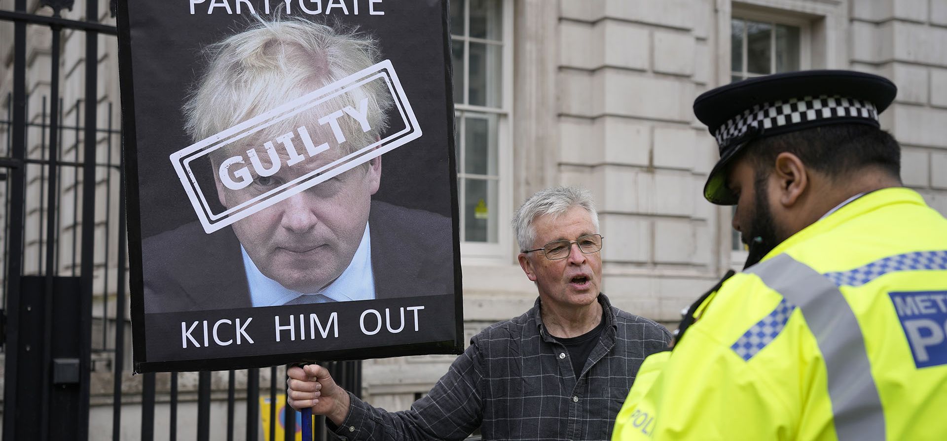 Un oficial de policía habla con un manifestante que sostiene un cartel que muestra al primer ministro británico, Boris Johnson, frente a la entrada de Downing Street en Londres, el miércoles 13 de abril de 2022. Johnson fue multado por violar las regulaciones de COVID-19 luego de acusaciones de fiestas de encierro en las oficinas del gobierno.