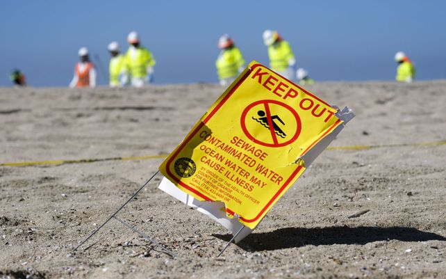 Una señal de advertencia para bañistas en la arena.Trabajadores con trajes protectores limpian la playa contaminada después de un derrame de petróleo en Huntington Beach, California.