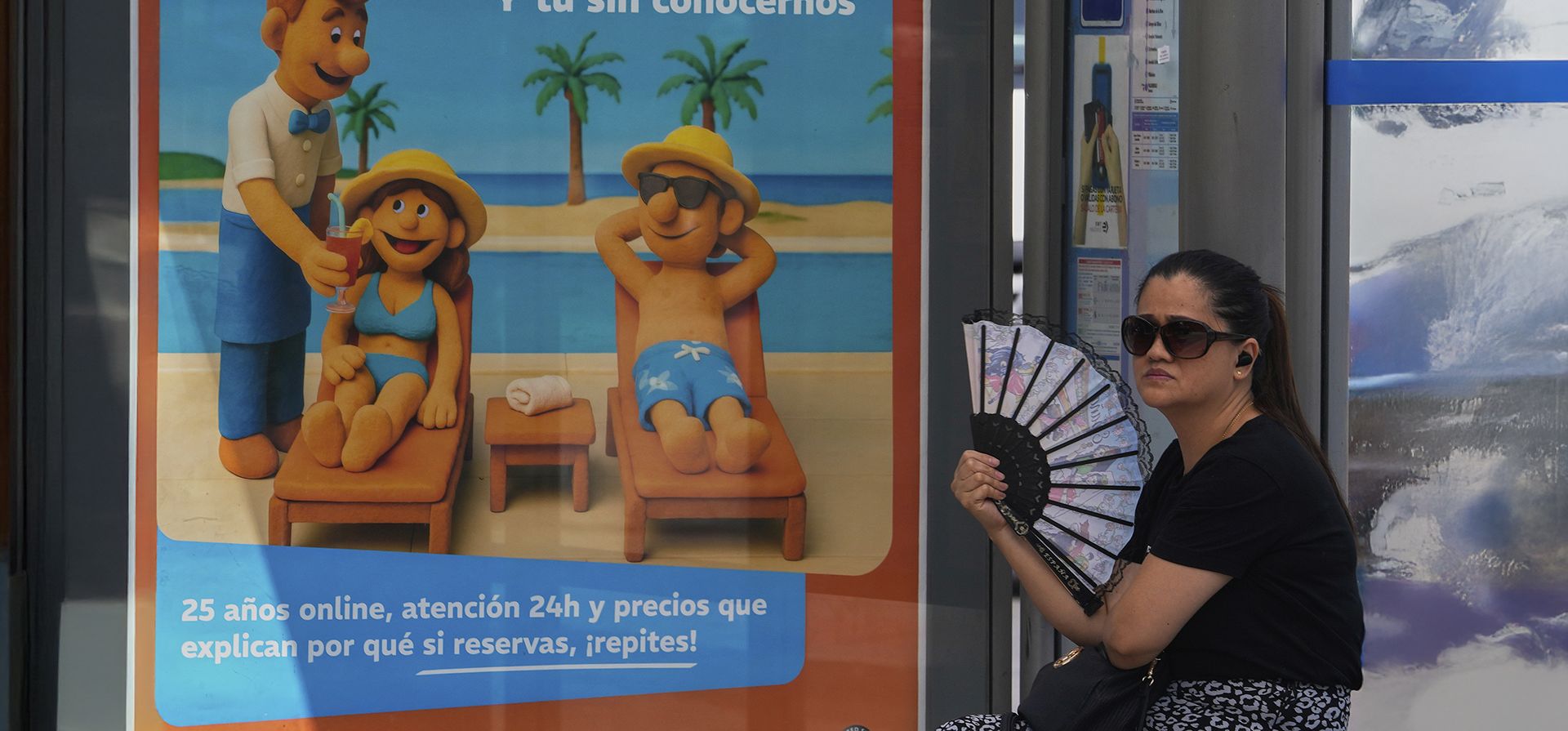 Una mujer se abanica en una parada de autobús, en un día caluroso, junto a un cartel de una agencia de viajes que muestra a gente en la playa en Madrid, España, el viernes 8 de agosto de 2025. La Comunidad de Madrid sigue en aviso naranja, después de varios días soportando altas temperaturas. (Foto AP/Paul White)