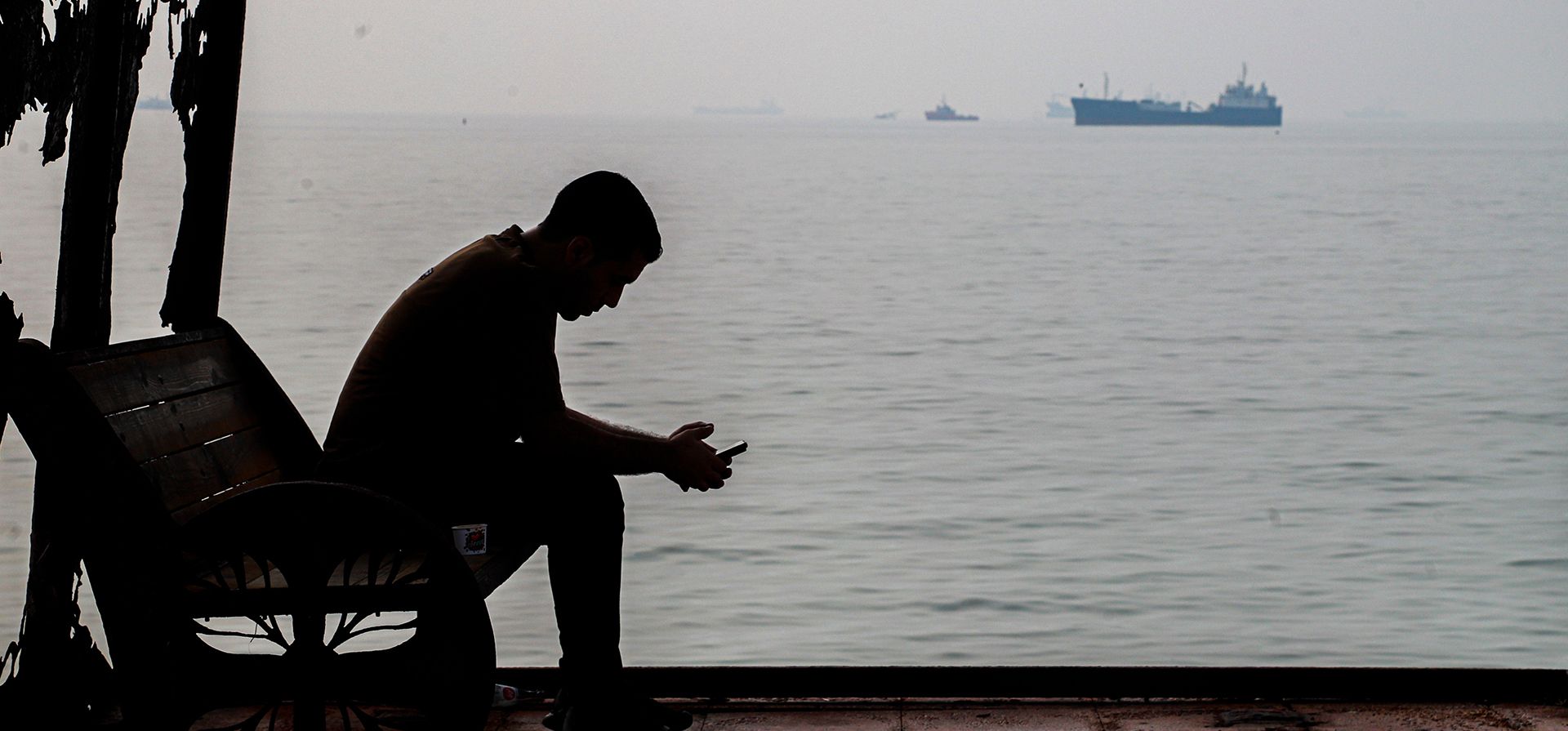 Un hombre mira su celular en un refugio junto a la costa mientras se ven barcos mercantes fondeados, en el estrecho de Ormuz, cerca de Bandar Abbas, Irán, el lunes 6 de octubre de 2026. (Amirhosein Khorgooi/ISNA via AP)  