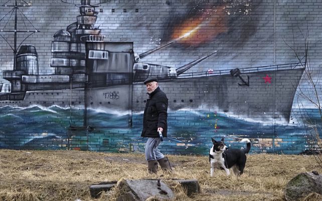 Un hombre camina con su perro junto a un graffiti en la pared de un edificio que representa un buque de guerra soviético de la Segunda Guerra Mundial en San Petersburgo