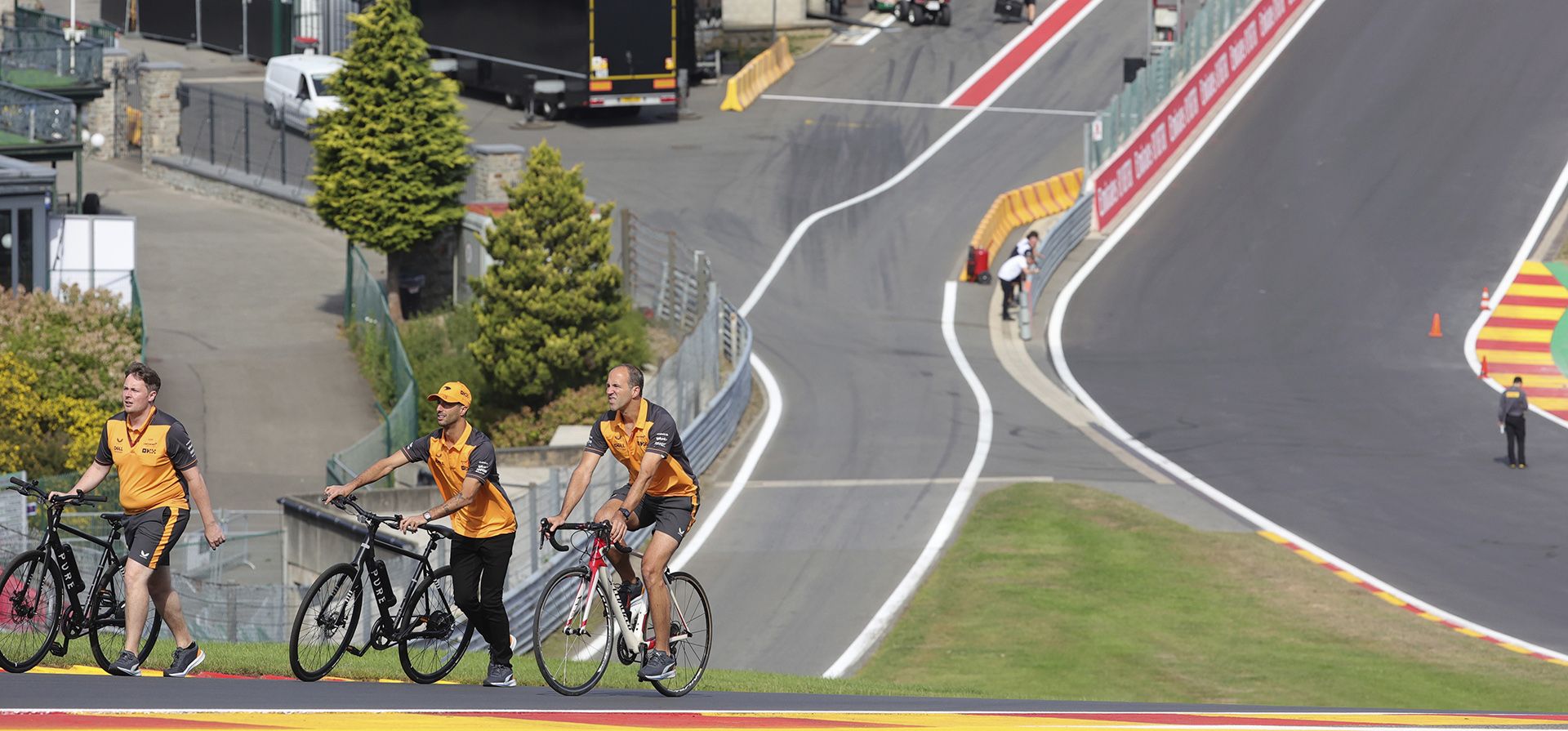 El piloto de McLaren Daniel Ricciardo de Australia, segundo a la izquierda, empuja una bicicleta en la pista con miembros de su equipo antes del Gran Premio de Fórmula Uno en el circuito de Spa-Francorchamps en Spa, Bélgica, el jueves 25 de agosto de 2022.