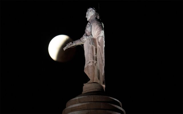 La sombra de la tierra cubre la luna llena durante un eclipse lunar parcial visible cerca de una estatua de George Washington en lo alto del Monumento en Baltimore, el viernes 19 de noviembre de 2021. El evento celeste, que ocurrió cuando la luna fue oscurecida por la sombra de la Tierra, durará poco más de seis horas, el eclipse lunar parcial de mayor duración en 580 años.