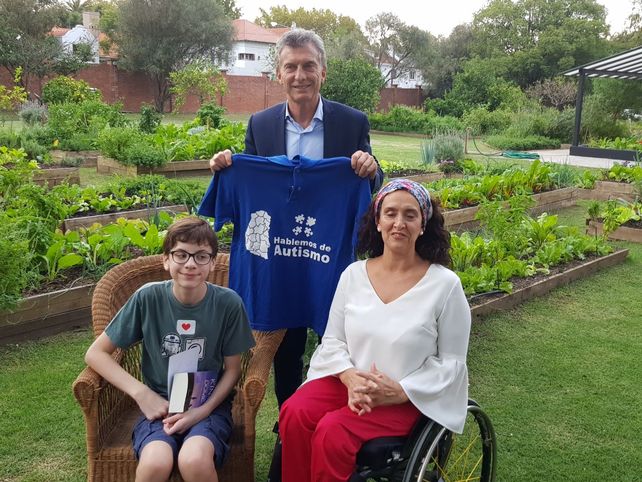 Santino sentado junto a la vicepresidente Gabriel Michetti. Macri sostiene la remera con el slogan: hablemos de autismo. De fondo