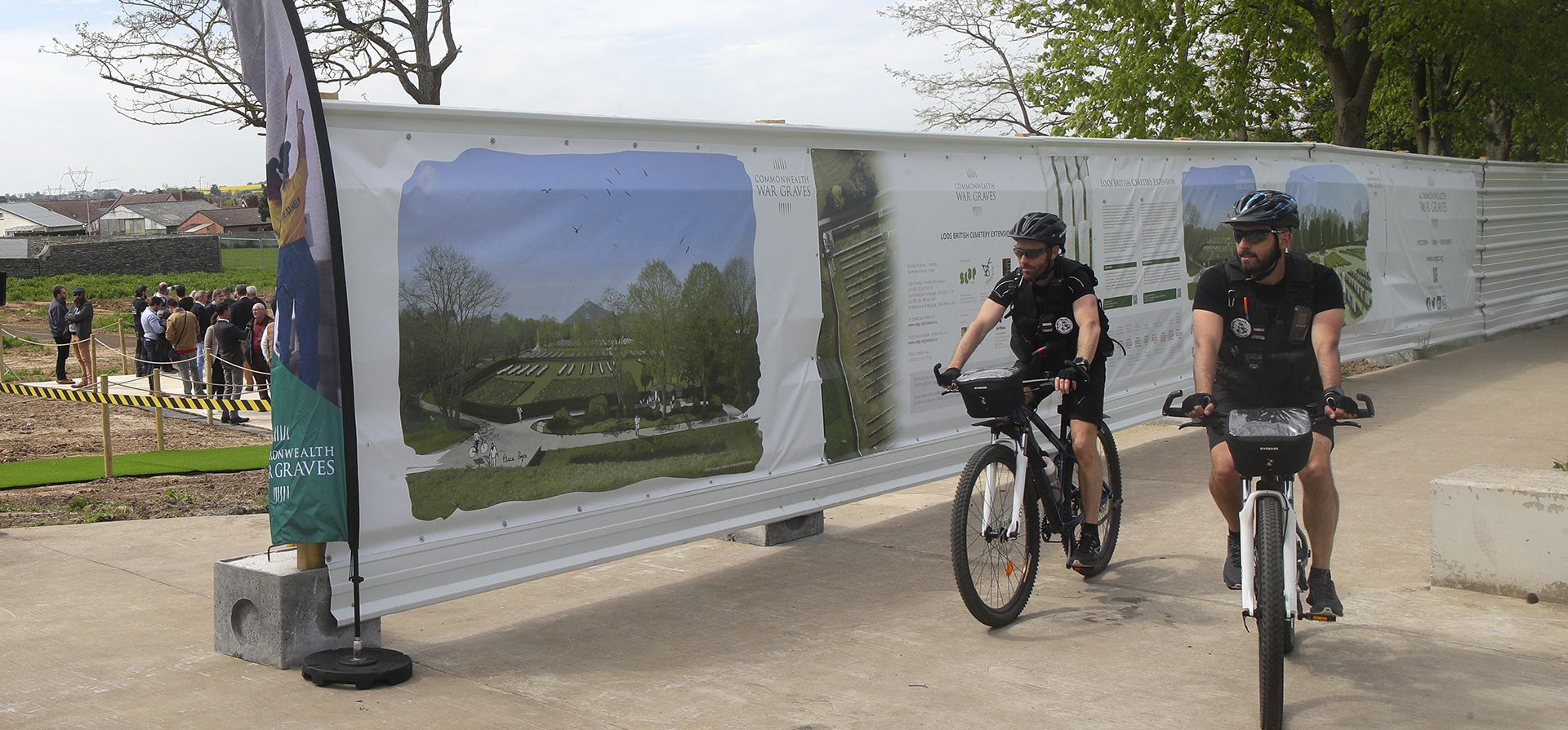 La policía en bicicleta pasa frente a una pancarta informativa antes de una ceremonia de colocación de cimientos en el Cementerio Británico de Loos en Loos-en-Gohelle, Francia, el jueves 4 de mayo de 2023. La Comisión de Tumbas de Guerra de la Commonwealth está ampliando el Cementerio Británico de Loos para proporcionar espacio de entierro para hasta a 1.200 víctimas de la Primera Guerra Mundial. (Foto AP/Michel Spingler)
