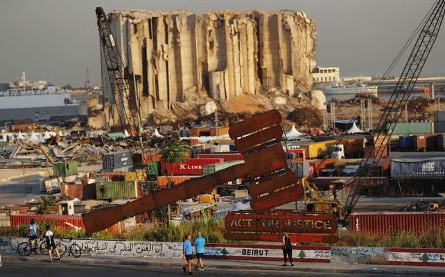Transeuntes pasan junto a un monumento con el símbolo de la justicia que se encuentra frente a imponentes silos de granos destruidos en la explosión masiva de agosto de 2020 en el puerto de Beirut que se cobró la vida de más de 200 personas.