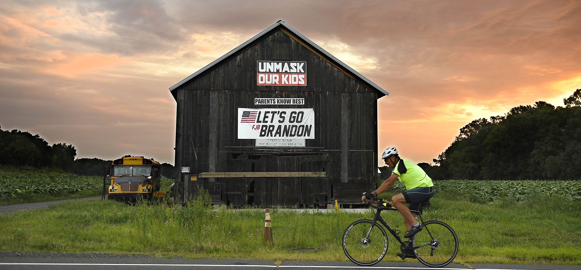Un ciclista pasa frente a un granero con una pancarta política mientras sale el sol el día de las elecciones primarias, el martes 9 de agosto de 2022, en Suffield, Connecticut.
