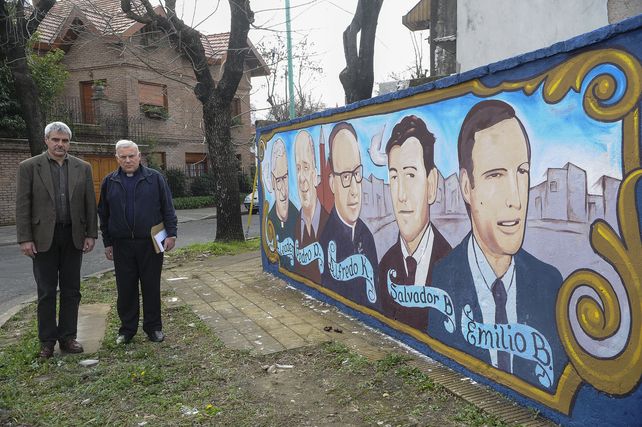 El recuerdo. Rolando Savino (izq.) y el sacerdote Rodolfo Capalozza al lado del mural con los rostros de los religiosos asesinados en el porteño barrio Belgrano.