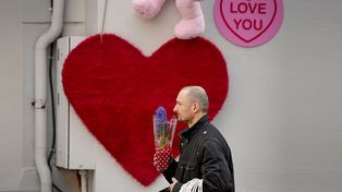 Un hombre que sostiene una maceta con una flor camina junto a una figura en forma de corazón en Bucarest, Rumania, el martes 14 de febrero de 2023.&nbsp; (Foto AP/Andreea Alexandru)