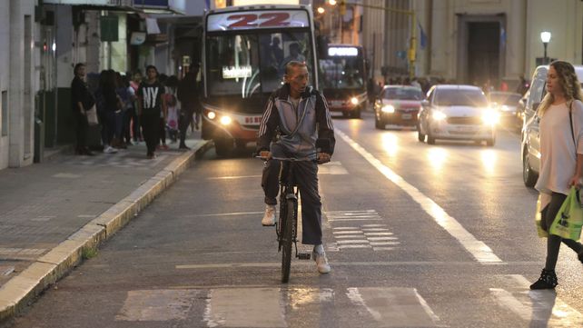 El ciclista espera que cruce la mujer mientras se vienen los autos y colectivos.&nbsp;
